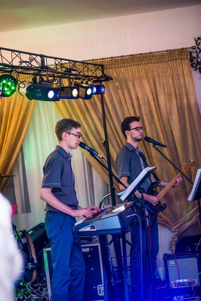 Dwóch muzyków w szarych koszulach gra na keyboardzie i gitarze elektrycznej na scenie z oświetleniem i złotymi zasłonami w tle.