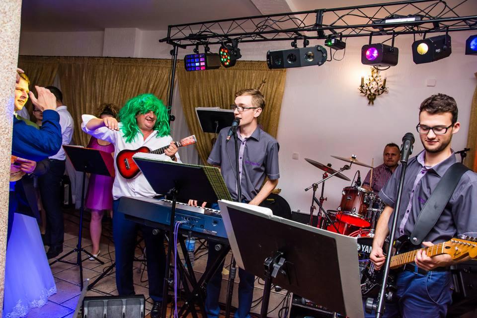 Muzycy grający na weselu: klawiszowiec w zielonej peruce gra na ukulele, wokalista śpiewa do mikrofonu, gitarzysta gra na gitarze elektrycznej, perkusista gra na perkusji.