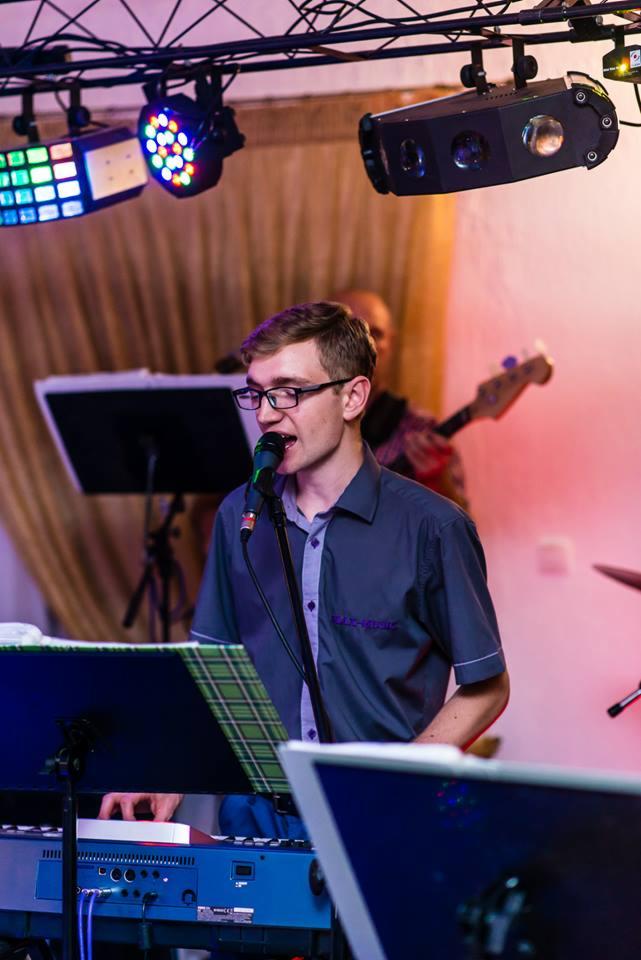 Młody mężczyzna w okularach śpiewa do mikrofonu grając na keyboardzie, w tle basista i oświetlenie sceniczne.