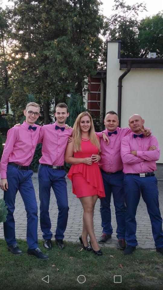 Pięcioosobowa grupa, w tym jedna kobieta w czerwonej sukience, pozuje na zewnątrz; mężczyźni ubrani w różowe koszule z muchami i niebieskie spodnie.