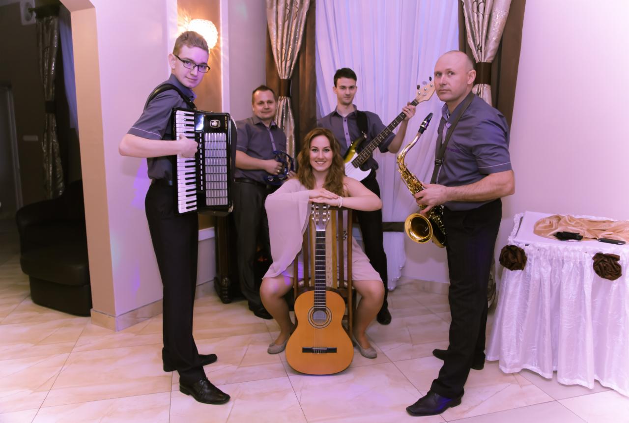 Zespół muzyczny w szarych koszulach prezentuje się z akordeonem, gitarą, saksofonem i gitarą basową, z wokalistką siedzącą na krześle z gitarą akustyczną na kolanach, w pomieszczeniu z beżowymi...