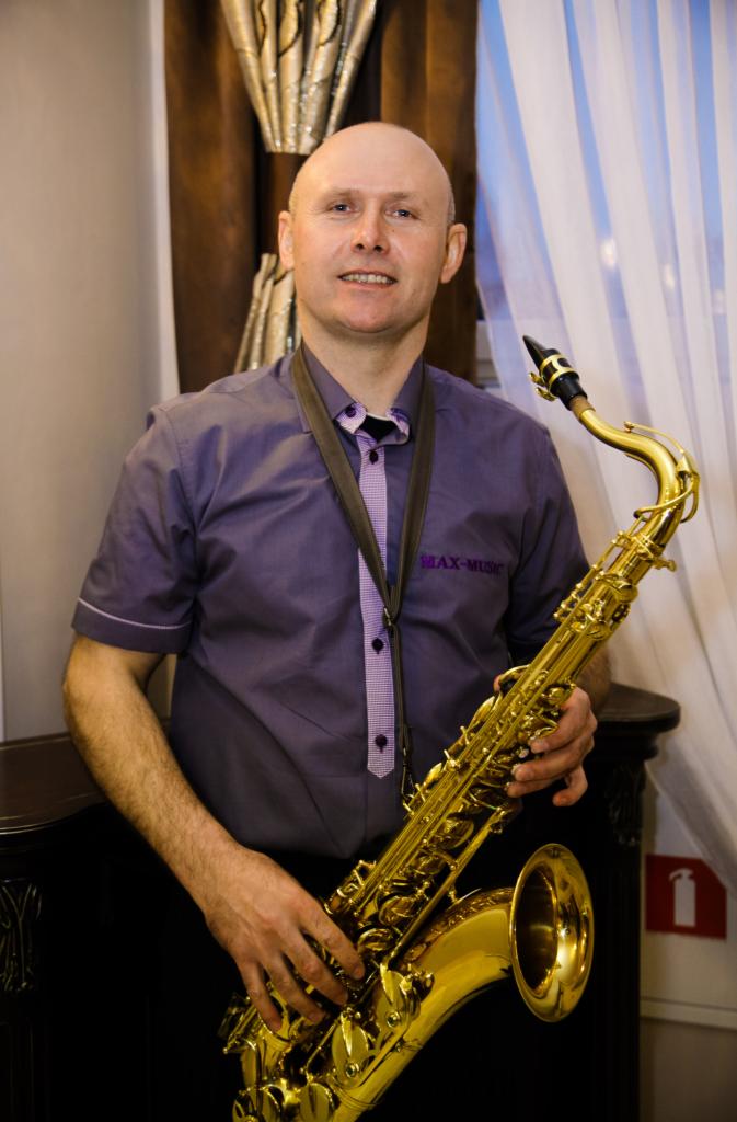 Portret mężczyzny z saksofonem, ubrany w fioletową koszulę z napisem MAX-MUSIC, na tle zasłon w odcieniach brązu i bieli.