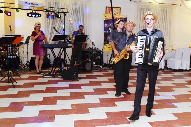 Występ zespołu na żywo: wokalistka z mikrofonem, klawiszowiec, saksofonista i akordeonista w pasujących kapeluszach na scenie z instrumentami.