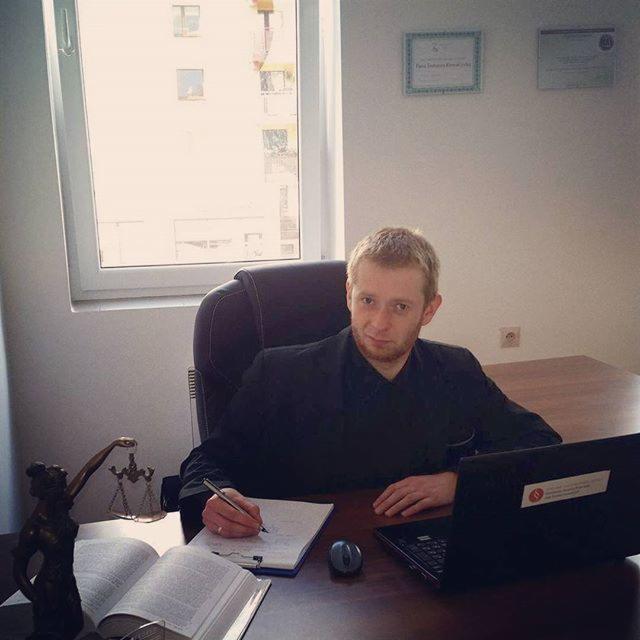 Młody mężczyzna w czarnym garniturze siedzi przy biurku z wagą Temidy, otwartą książką, notatnikiem, długopisem i laptopem, pisząc coś w notatniku. Na ścianie wiszą dwa certyfikaty.