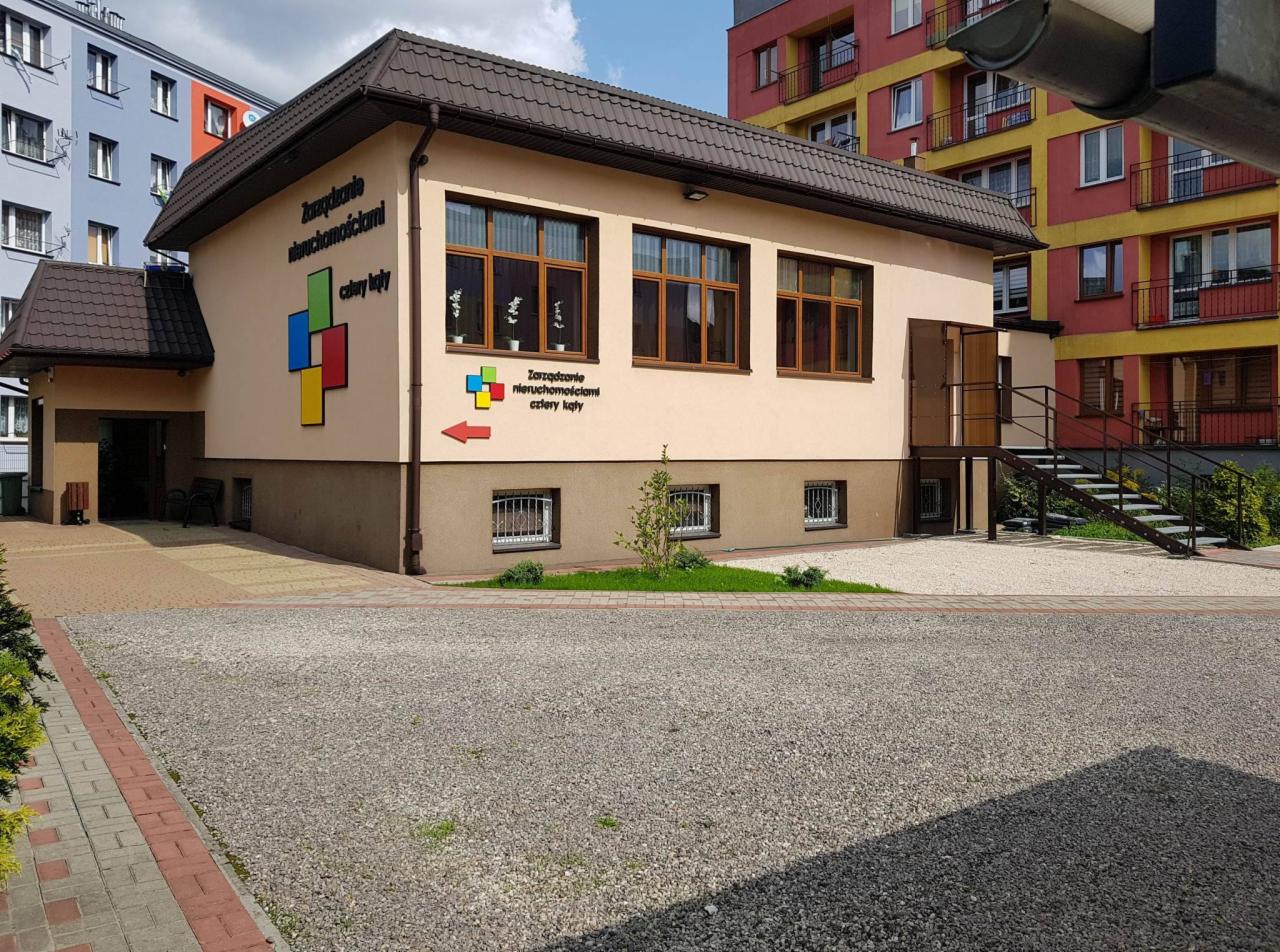 Budynek biurowy z napisem 'Zarządzanie Nieruchomościami cztery kąty' i kolorowym logo, widoczne schody wejściowe i żwirowy parking.