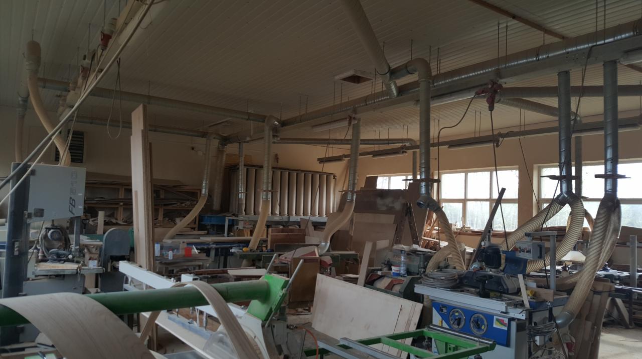 Wnętrze warsztatu stolarskiego z maszynami do obróbki drewna i systemem odsysania pyłu, widoczne płyty drewniane i elementy konstrukcyjne w trakcie produkcji.