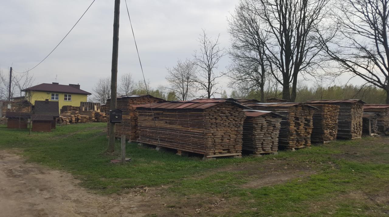 Składowane pakiety tarcicy na zewnątrz, zabezpieczone blachą przed deszczem, widoczne drewniane deski i elementy konstrukcyjne, w tle budynek mieszkalny i drzewa bez liści.