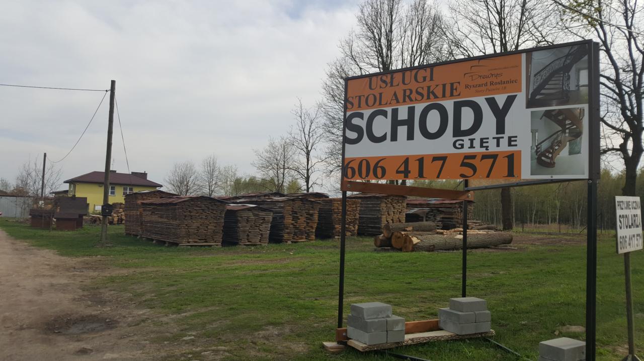 Tablica reklamowa usług stolarskich z numerem telefonu na tle składowanego drewna i żółtego domu.