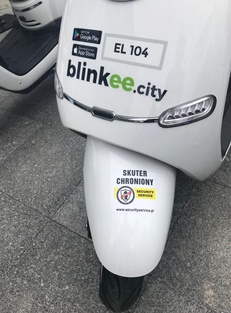 Biały skuter Blinkee.city z naklejką 'Skuter Chroniony Security Service' na przednim błotniku.