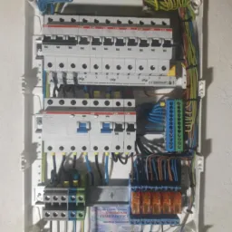 Rozdzielnica elektryczna z zainstalowanymi bezpiecznikami, widoczne okablowanie i listwy zaciskowe, naklejka z danymi firmy elektrycznej w dolnej części.