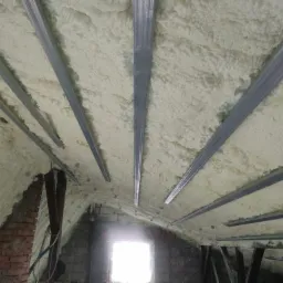 Poddasze z widoczną pianką izolacyjną między metalowymi profilami, częściowo otynkowane ściany z cegły i pustaków, okno w tle.