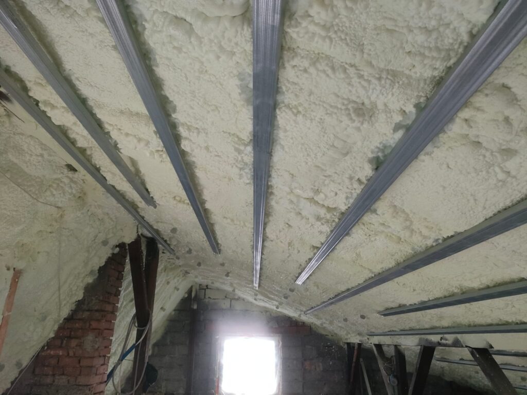 Poddasze z widoczną pianką izolacyjną między metalowymi profilami, częściowo otynkowane ściany z cegły i pustaków, okno w tle.