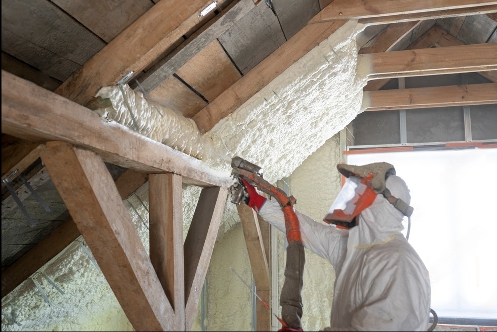 Pracownik w kombinezonie ochronnym aplikuje pianę izolacyjną na drewnianą konstrukcję poddasza, widoczne nierównomierne pokrycie i struktura piany.