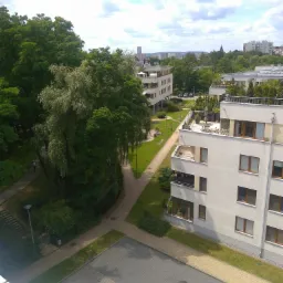 Widok z góry na osiedle mieszkaniowe z zielenią, ścieżkami spacerowymi i parkingiem, w tle panorama miasta pod błękitnym niebem z chmurami.
