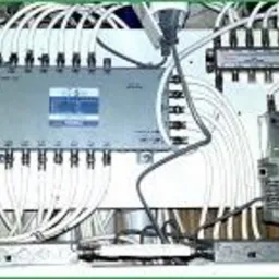 Rozdzielnica telekomunikacyjna z licznymi kablami koncentrycznymi, widoczne złącza F i elementy aktywne, całość zamontowana na białej płycie.