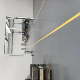 Szara podłoga w hali przemysłowej z wyznaczoną żółtą linią komunikacyjną, widoczne palety i otwarte drzwi do magazynu.