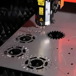 Proces wycinania laserowego w metalowej płycie, tworzący otwory o skomplikowanych, zębatych kształtach. Widoczna głowica lasera z oznaczeniami bezpieczeństwa, skierowana na obrabiany materiał.