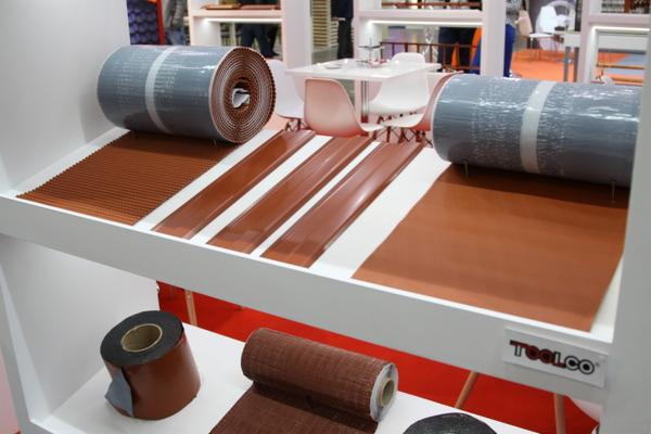Ekspozycja materiałów dachowych w kolorze ceglastym, w tym rolek papy i pasów blachy na białym stojaku, z widocznym logo Toolco.