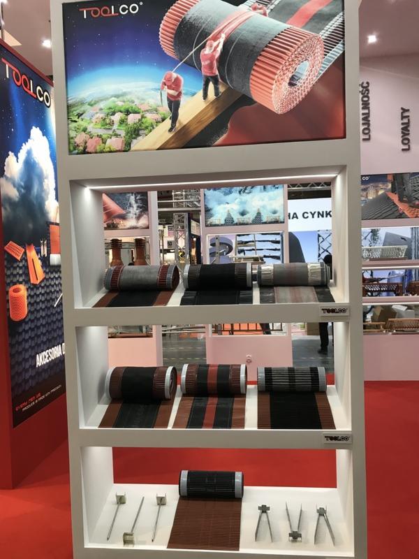 Ekspozycja materiałów dekarskich na targach: rolki papy w różnych kolorach i wzorach na białych półkach, narzędzia dekarskie na dolnej półce, w tle grafika z pracownikami na dachu i logo firmy.