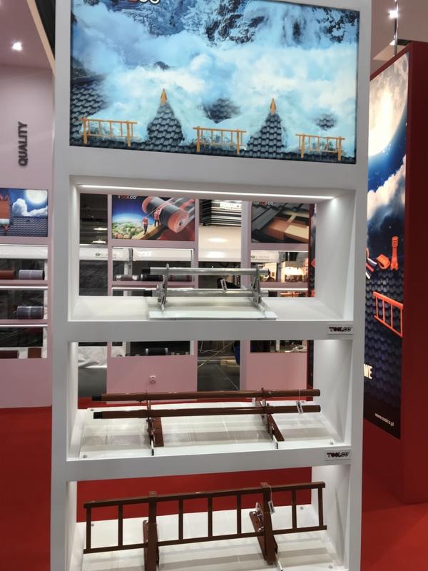 Ekspozycja materiałów dekarskich na targach: metalowe elementy do montażu na dachu zaprezentowane na białym regale z podświetleniem, w tle grafika imitująca zimowy krajobraz górski z pokrytymi...