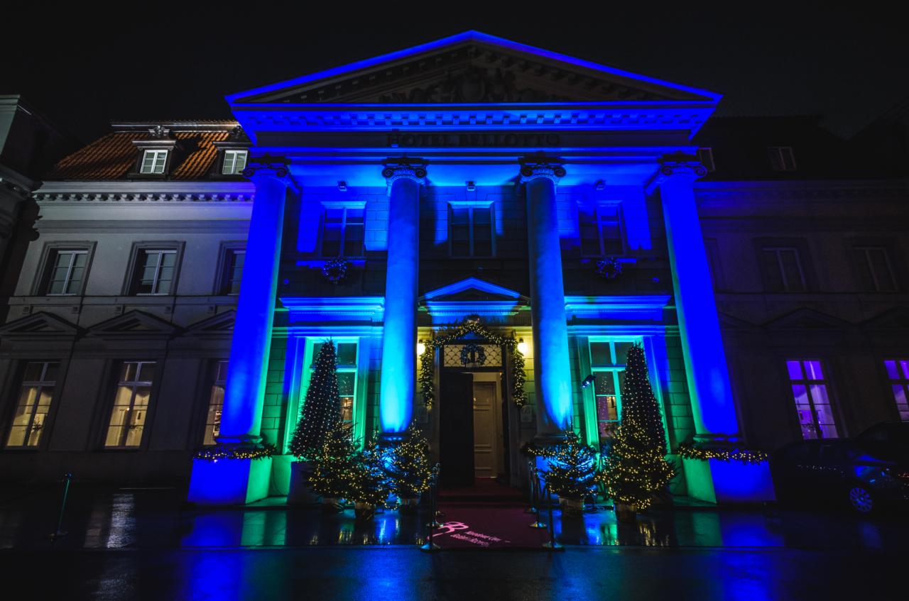 Elegancki budynek z kolumnami, oświetlony na niebiesko i zielono, udekorowany choinkami przed wejściem, nocne ujęcie.