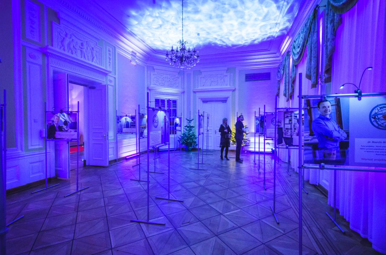 Wnętrze sali balowej z ekspozycją fotografii i oświetleniem w odcieniach niebieskiego, para gości w tle, choinka, żyrandol.