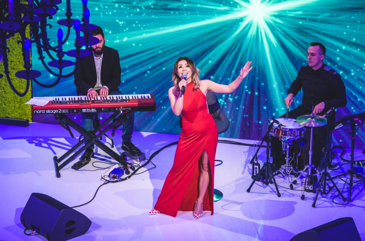 Wokalistka w czerwonej sukni śpiewa na scenie podczas występu zespołu muzycznego, z klawiszowcem i perkusistą, na tle dekoracji z efektem gwiazd.