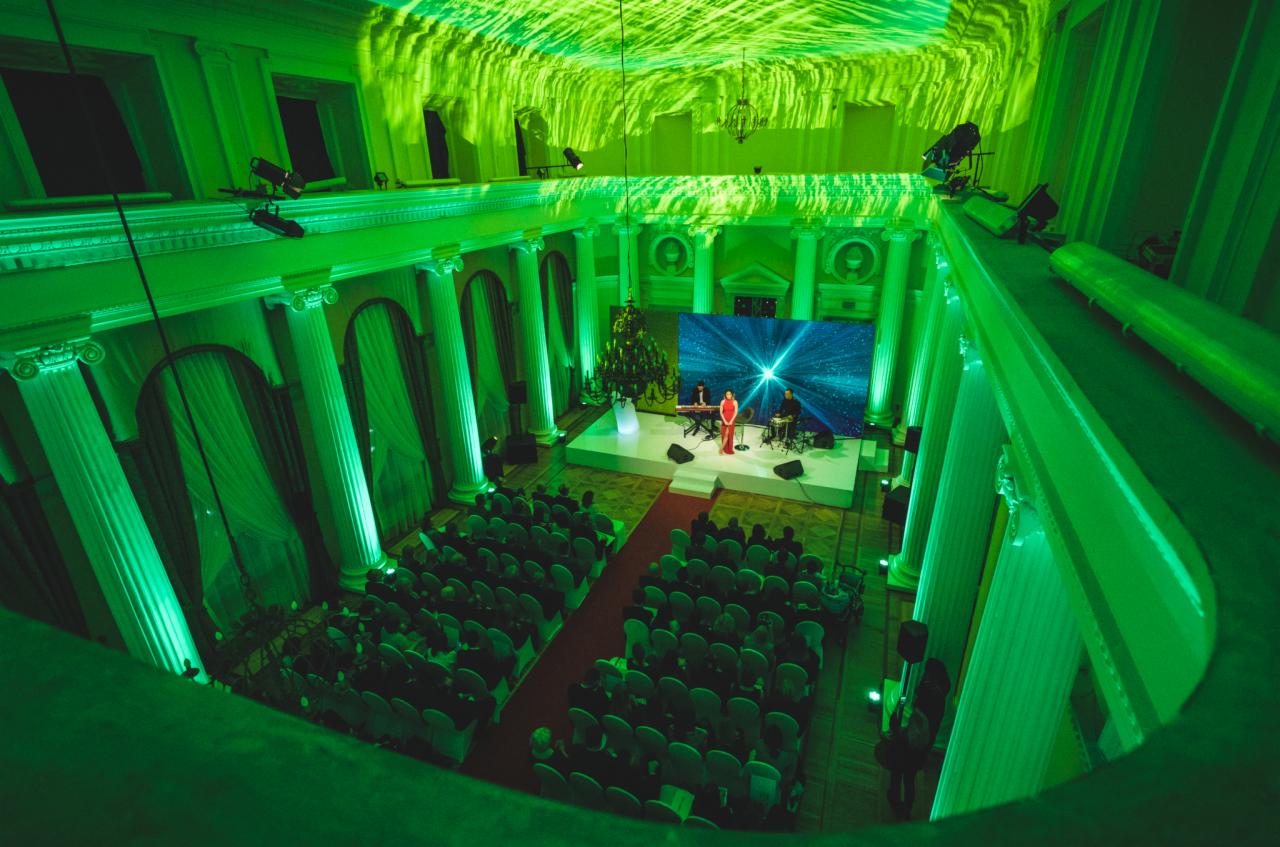 Widok z góry na salę balową z kolumnami oświetloną zielonym światłem, z publicznością siedzącą na krzesłach i zespołem muzycznym występującym na scenie przed dużym ekranem LED wyświetlającym motyw...