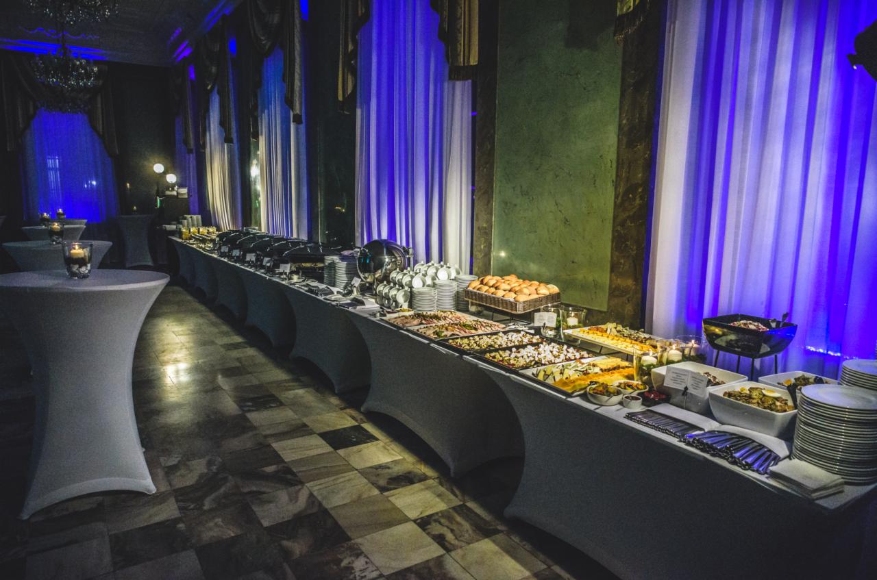 Długi stół bufetowy z różnorodnymi daniami, talerzami i sztućcami w eleganckiej sali z marmurową podłogą, oświetlony niebieskim światłem i dekorowany białymi zasłonami.