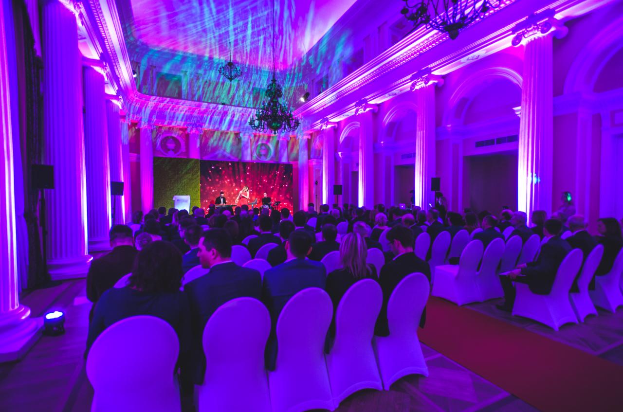 Duża sala balowa z rzędami białych krzeseł wypełnionych publicznością, oświetlona fioletowymi światłami i z dekoracjami świetlnymi na suficie oraz sceną z zieloną ścianą w tle.