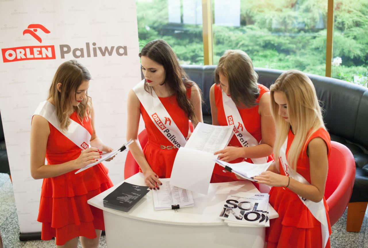 Cztery hostessy w czerwonych sukienkach i szarfach Orlen Paliwa pracują przy okrągłym białym stole, przeglądając dokumenty i notatki, z banerem reklamowym w tle i widokiem na zieleń za oknem.