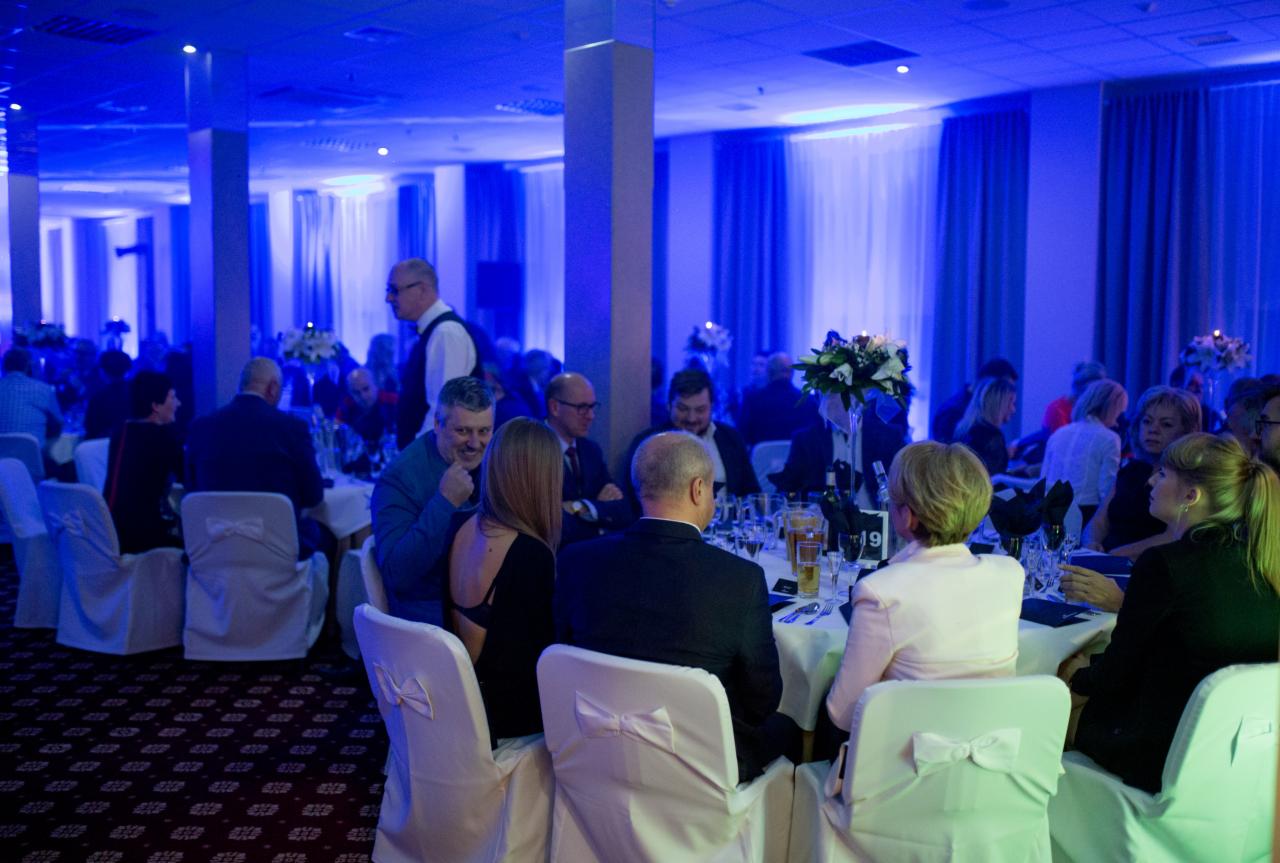 Uczestnicy siedzą przy okrągłych stołach podczas oficjalnego wydarzenia oświetlonego niebieskim światłem, białe krzesła z kokardami, dekoracje kwiatowe i napoje na stołach.