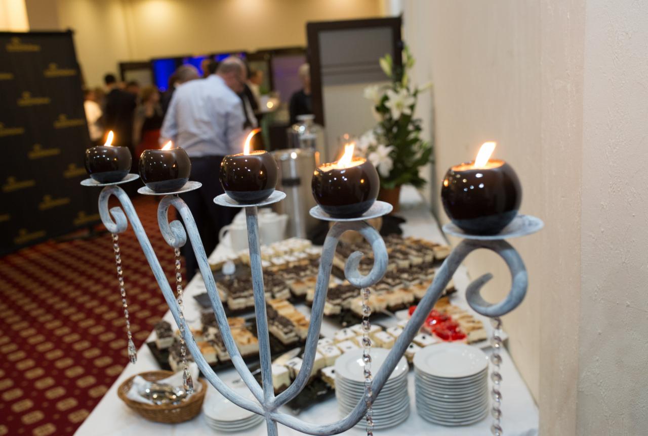 Dekoracyjny świecznik z czterema płonącymi kulistymi świecami na tle stołu z deserami i gośćmi wydarzenia.