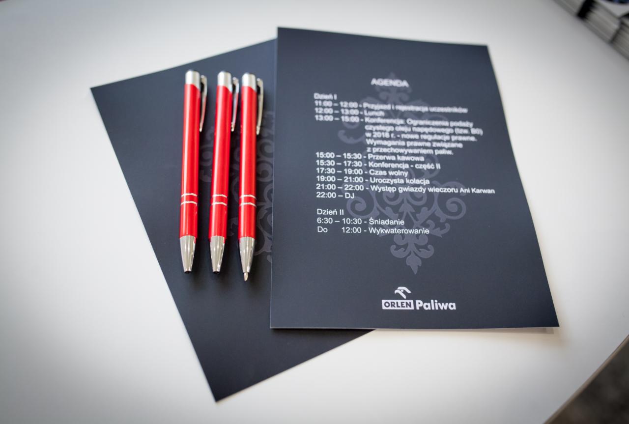 Trzy czerwone długopisy leżące obok czarnego programu konferencji Orlen Paliwa, widoczne godziny poszczególnych wydarzeń.