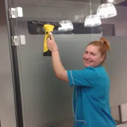 Uśmiechnięta kobieta w niebieskim uniformie czyści szklaną ścianę biurową za pomocą żółtej myjki do okien.