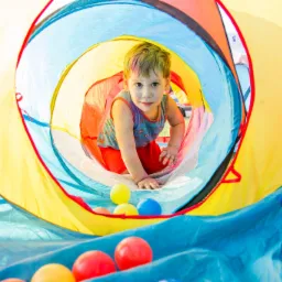 Uśmiechnięty chłopiec w tunelu z kolorowej tkaniny, otoczony plastikowymi piłeczkami.