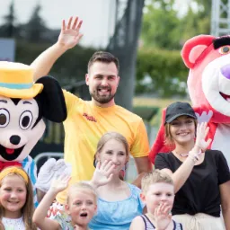 Animatorzy w strojach Myszki Miki i pluszowego misia oraz animator w żółtej koszulce pozują z grupą uśmiechniętych dzieci i nastolatków na tle zieleni.