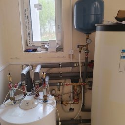Usługi instalatorskie Piotr Rynkowski - Biały bojler elektryczny z podłączeniami hydraulicznymi, rurami w izolacji i zbiornikiem wyrównawczym, zamontowany w pomieszczeniu z oknem.
