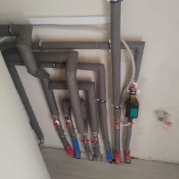 Szare, zaizolowane rury instalacji grzewczej z zaworami i pompą obiegową Wilo, zamontowane przy białej ścianie z widocznymi śladami po instalacji.