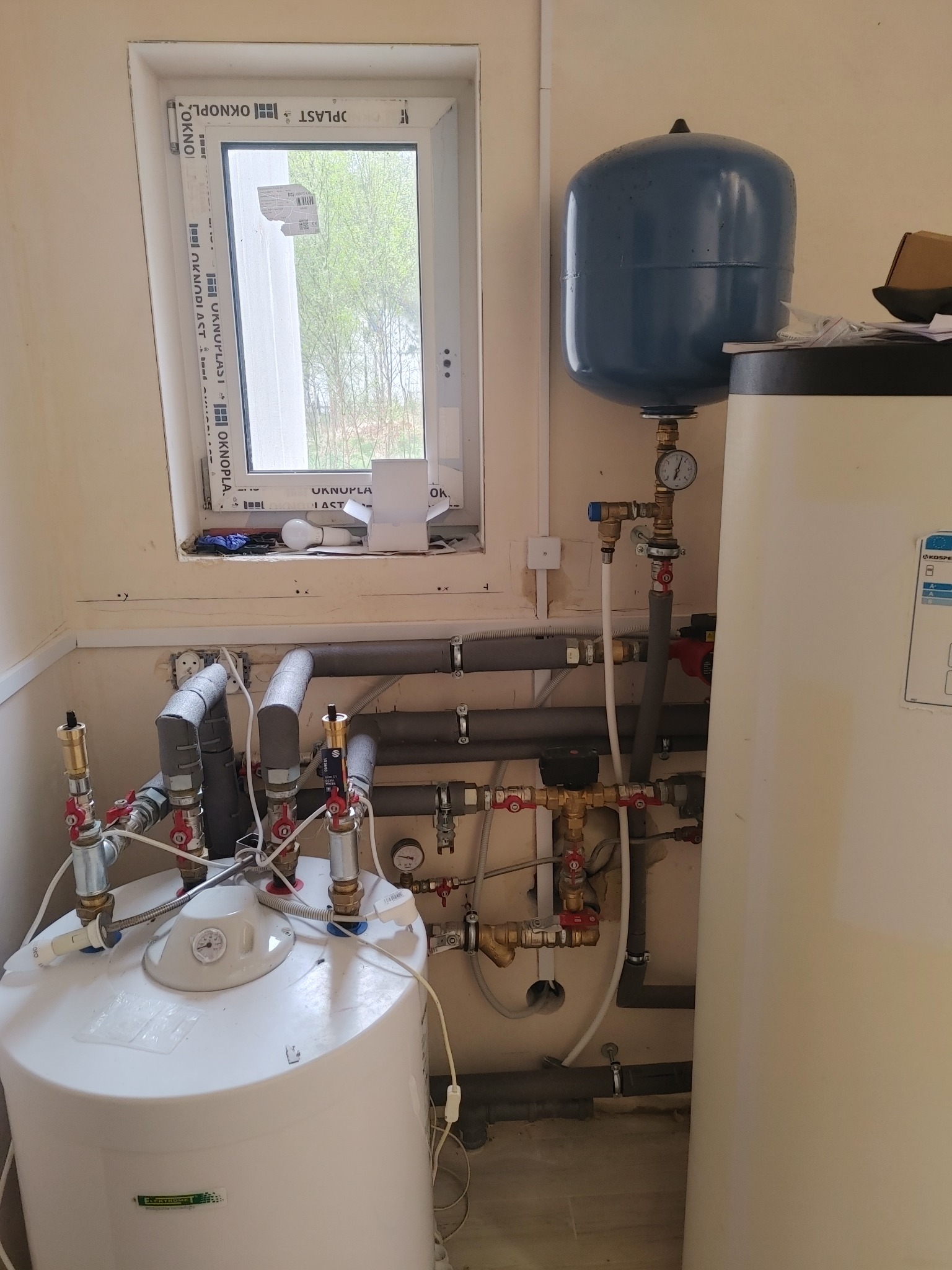 Biały bojler elektryczny z podłączeniami hydraulicznymi, rurami w izolacji i zbiornikiem wyrównawczym, zamontowany w pomieszczeniu z oknem.