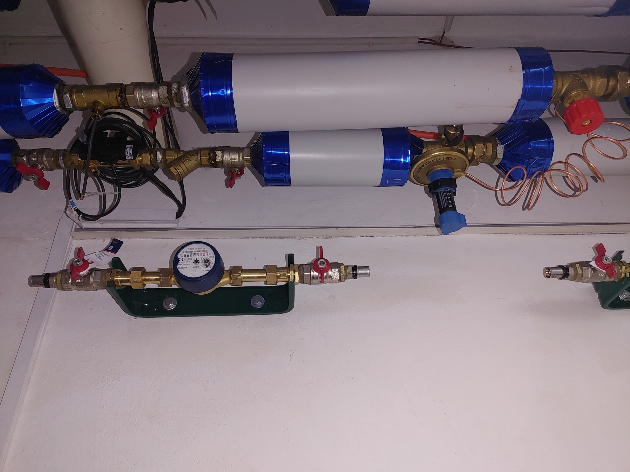 Instalacja wodna z filtrami cylindrycznymi owiniętymi niebieską folią, licznik wody z niebieską obudową, zawory z czerwonymi pokrętłami, miedziane rurki, zamontowane na białej ścianie.