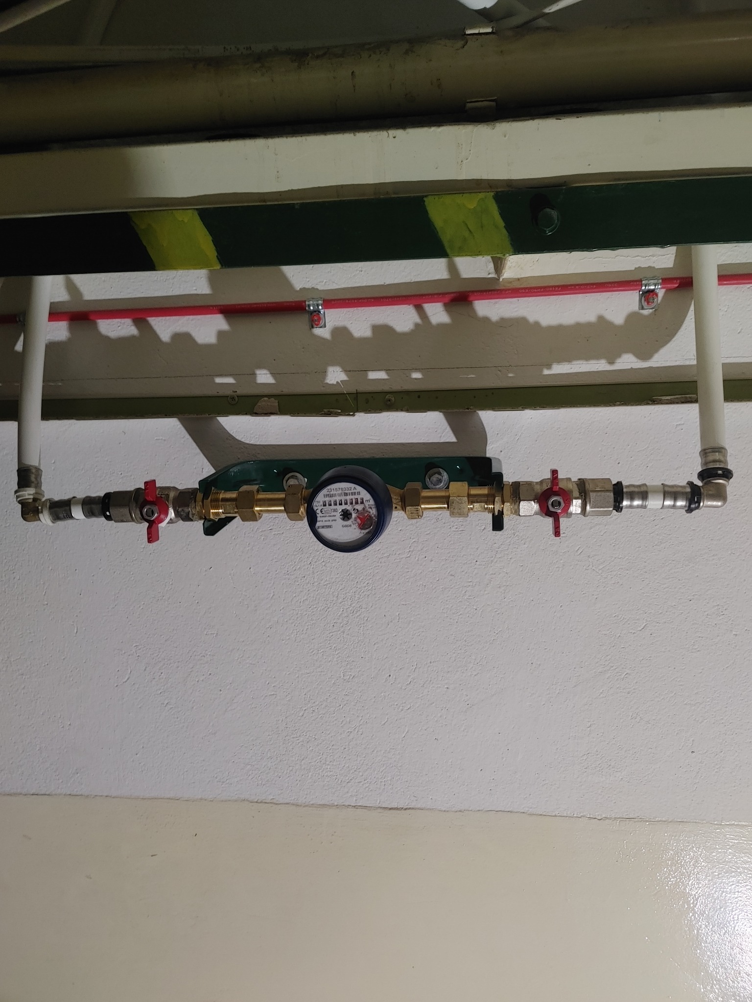 Zamontowany licznik wody z czerwonymi zaworami odcinającymi, przymocowany do ściany w Dobiesławiu, widoczne rury doprowadzające i odprowadzające wodę.