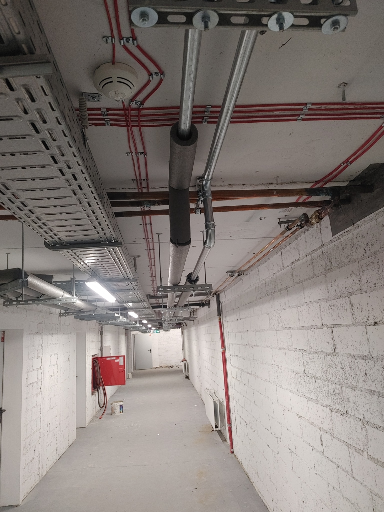 Instalacje w piwnicy budynku: rury grzewcze z izolacją, korytka kablowe pod sufitem, instalacja przeciwpożarowa z hydrantem wnękowym i czujnik dymu na suficie.