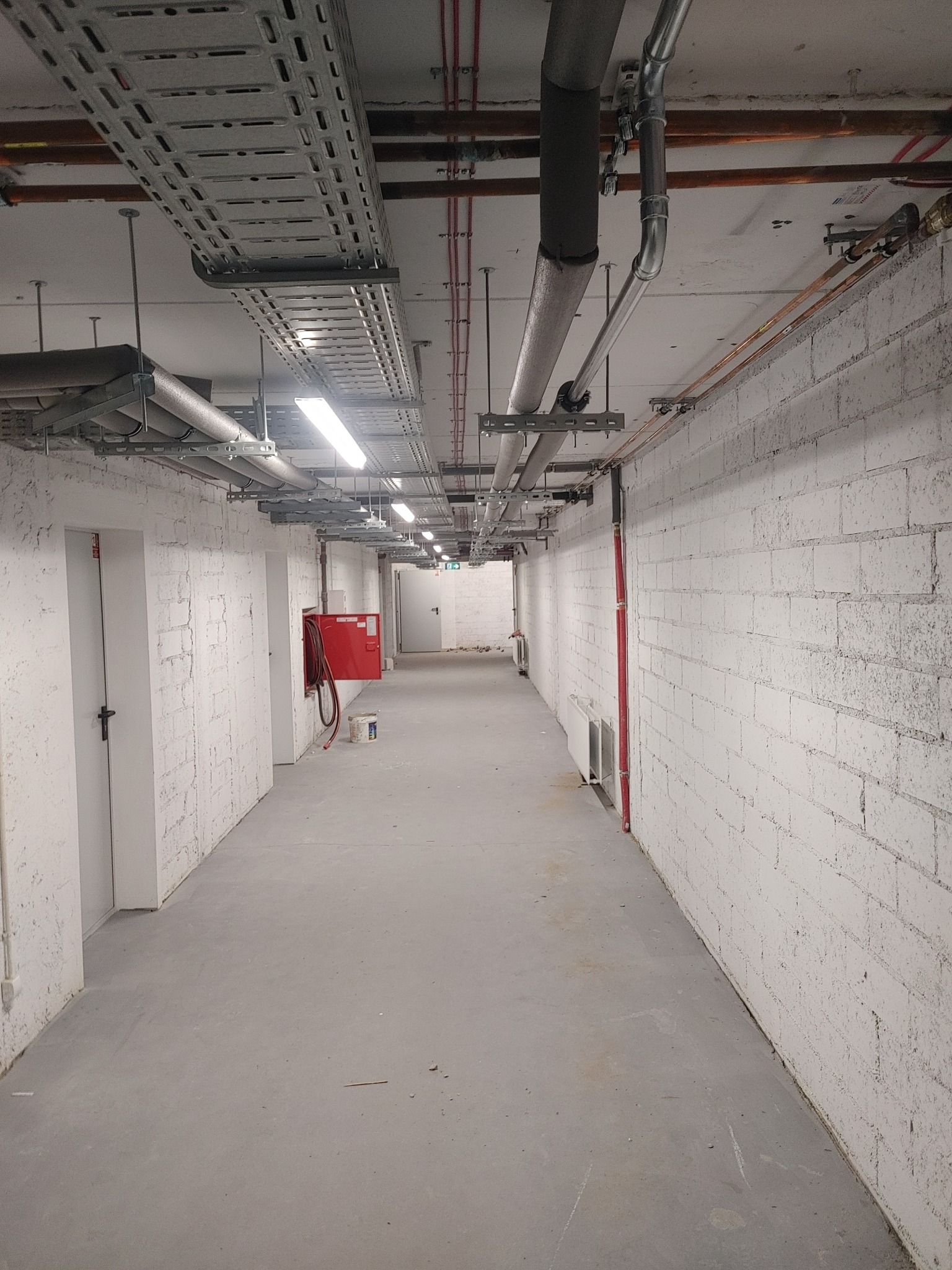 Korytarz piwniczny z instalacjami pod sufitem: rury miedziane, stalowe, koryta kablowe, instalacja przeciwpożarowa z czerwoną szafką hydrantową przy ścianie z białej cegły.
