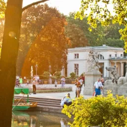 Łazienki Królewskie w Warszawie w słoneczny dzień, z widocznym Pałacem na Wodzie, rzeźbami, fontanną i spacerującymi ludźmi.