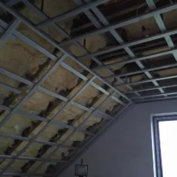 Skośne poddasze z widoczną konstrukcją metalową i ociepleniem z wełny mineralnej, okno w tle, widok z dołu.