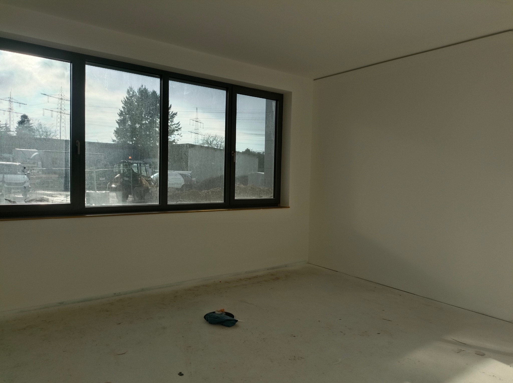 Jasne wnętrze po malowaniu, widoczne duże okno z widokiem na zewnątrz. Świeżo pomalowane ściany i sufit. Na podłodze widoczne ślady po pracach malarskich.