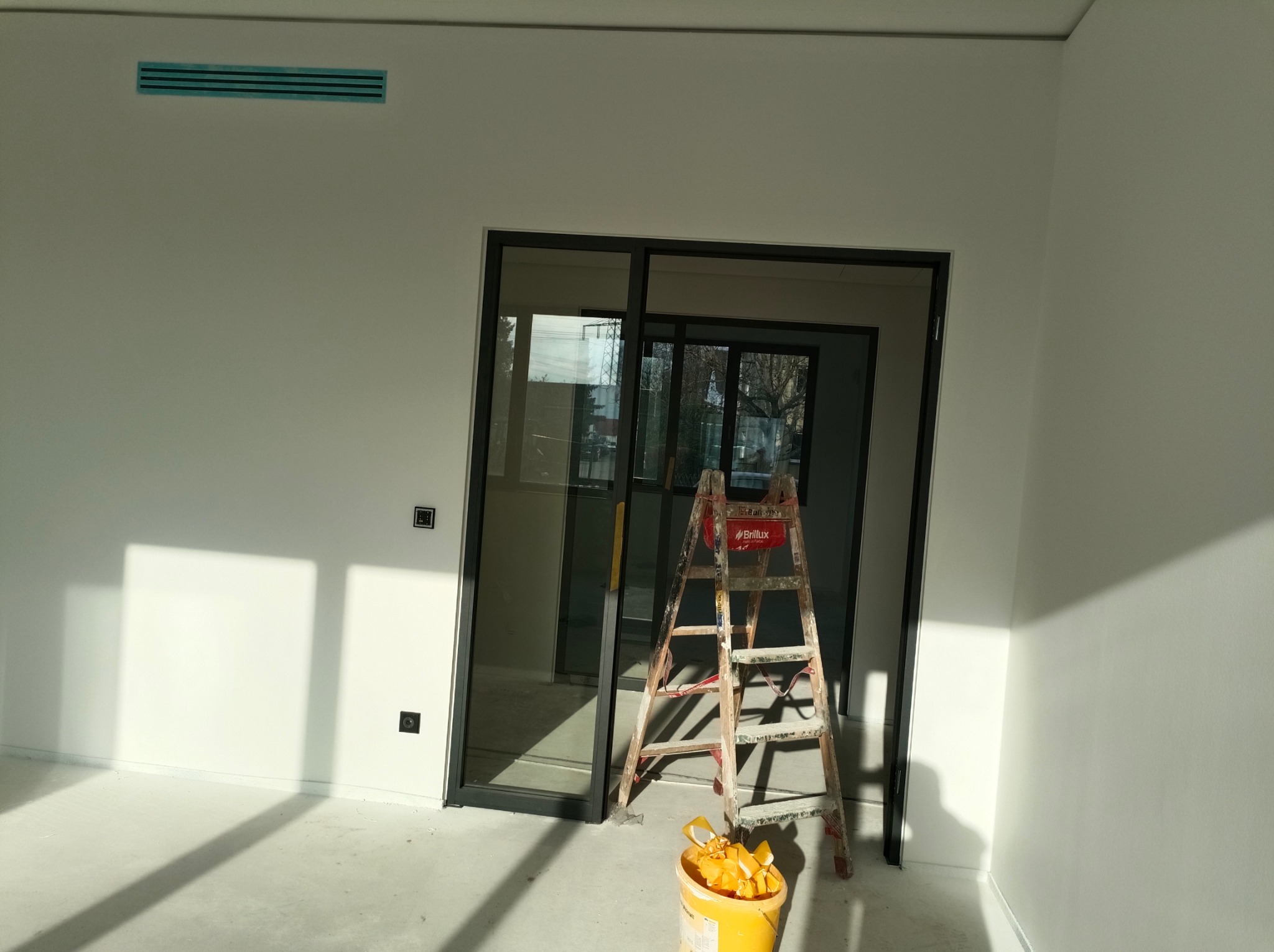 Białe, świeżo pomalowane wnętrze z drabiną i wiadrem farby przy czarnych, przesuwnych drzwiach. Widoczny fragment wentylacji. Minimalistyczny styl.