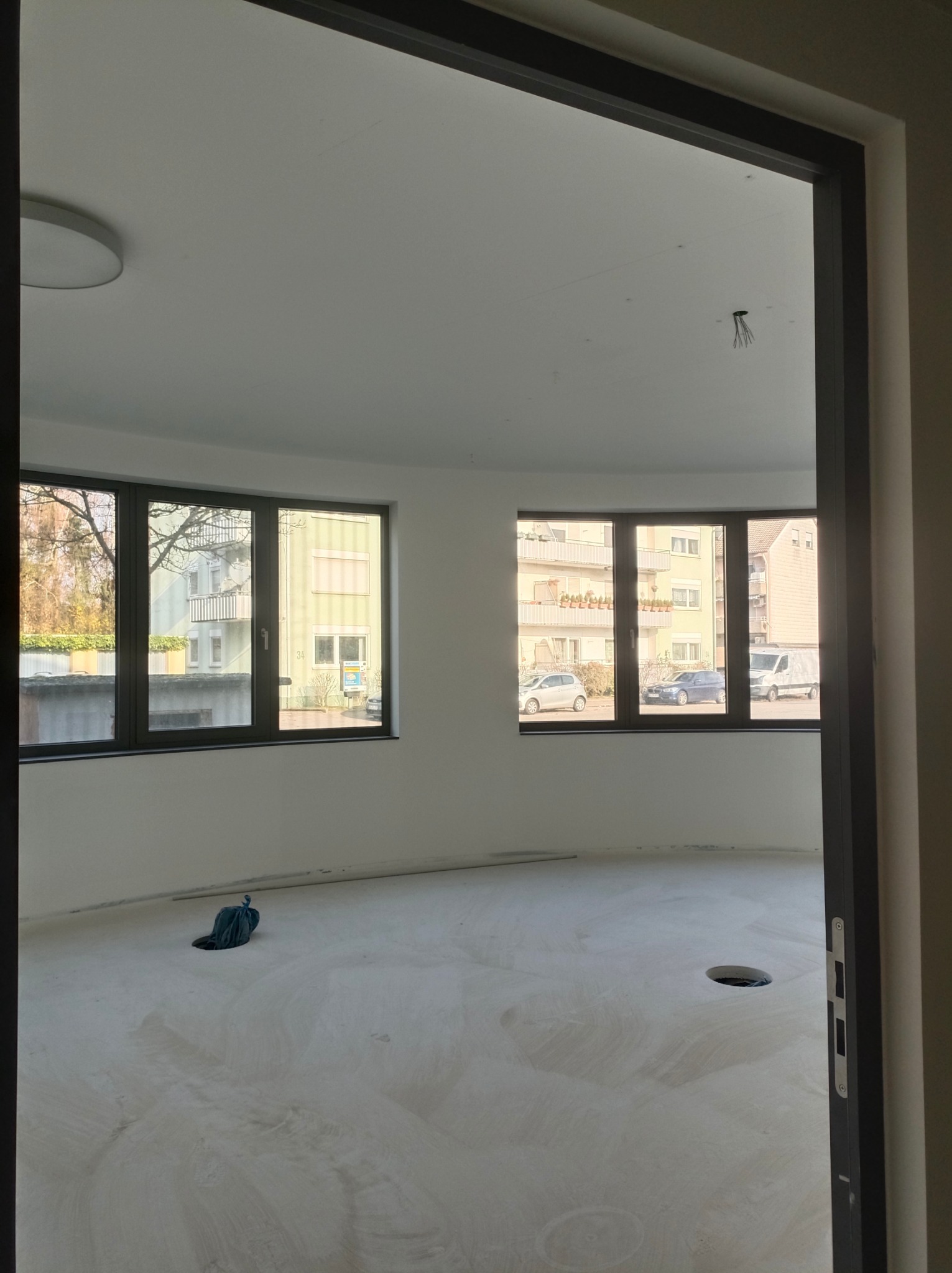 Wnętrze w trakcie prac wykończeniowych: surowa podłoga, świeżo pomalowane ściany, okna z widokiem na budynki. Widoczne ślady szpachlowania na podłodze.