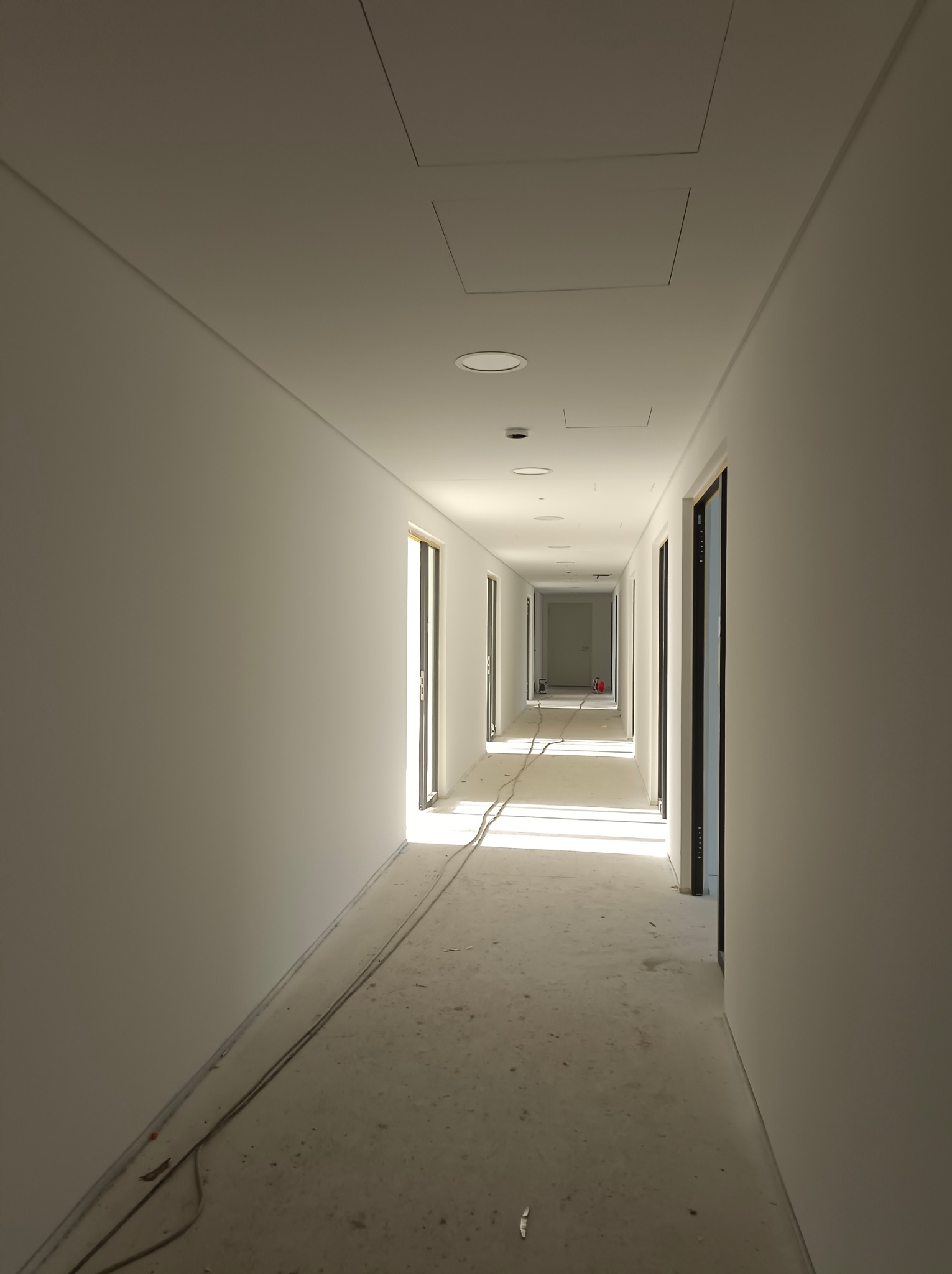 Długi korytarz w trakcie wykańczania, świeżo pomalowane ściany na biało, widoczne kable na podłodze i otwory rewizyjne w suficie. Perspektywa zbieżna.
